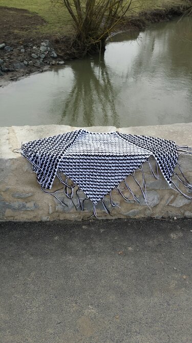 Schwarz-weißes Dreieckstuch mit Hahnentritt und Fransen auf einer grauen Steinmauer vor einem Fluss und Bäumen.