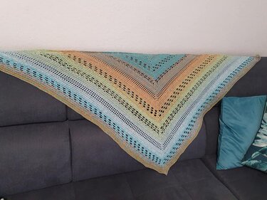 Handgehäkeltes Dreieckstuch mit Farbverlauf und Lochmuster auf grauem Sofa.