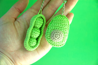 Hand holding two green crocheted keychains: a pea pod with three peas and an avocado half with a beige spiral pit, both on short metal chains