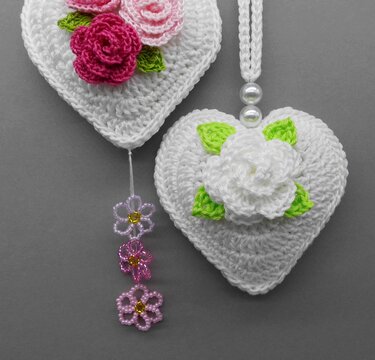 Zwei weiße gehäkelte Herzanhänger mit Rosenapplikationen, grünen Blättern und Perlen auf grauem Hintergrund