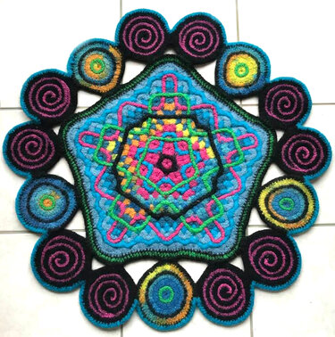 Gehäkeltes, farbenfrohes Pentagon mit spiralförmigen und konzentrischen Kreisornamenten auf hellem Untergrund.