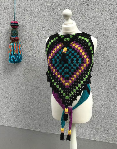 Gehäkeltes schwarzes Poncho-Top mit bunten Granny Squares und Quasten auf einer Schneiderpuppe vor grauer Wand.