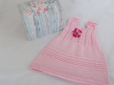 Rosa gestricktes Babykleid mit Zopfmuster und kleiner Blumenapplikation, liegend auf weißem Stoff neben einer dekorativen Holzbox.