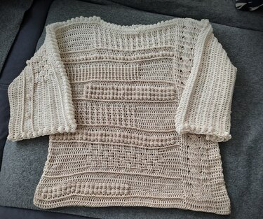 Beiger handgehäkelter Damenpullover mit Noppen-, Relief- und Zopfmustern und 3/4-Ärmeln, flach ausgelegt.