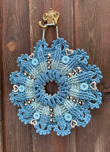 Gehäkelter runder Kranz in Blau und Hellblau mit Knöpfen, Glöckchen und Herzverzierungen, aufgehängt an einem verzierten Haken vor einer Holzwand.