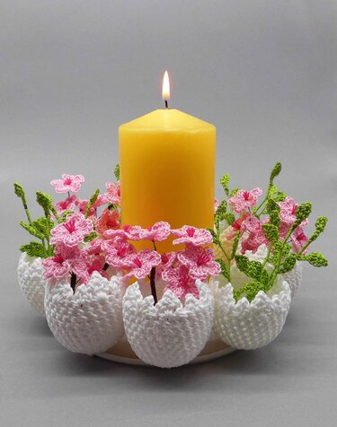 Lit yellow pillar candle surrounded by white crocheted eggshell-shaped holders, each filled with pink crocheted flowers and green stems, on a neutral background.
