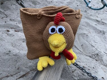 Verspielte gehäkelte Tasche mit großem Amigurumi-Hühnerkopf, gelbem Schnabel, rotem Kamm und gelben Füßen auf sandigem Boden