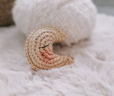 Nahaufnahme eines beigen, gehäkelten Amigurumi-Mondes mit geschlossenen Augen und rosa Wangen auf einem flauschigen hellen Untergrund.