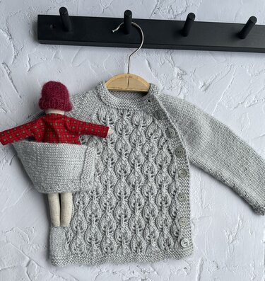 Gestrickter hellgrauer Babycardigan mit dekorativen Blattreihen, seitlicher Knopfleiste, Stoffpuppe in Tasche, aufgehängt an Holzbügel.