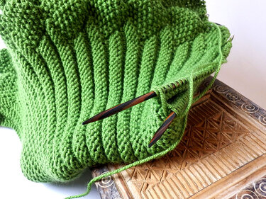 Nahaufnahme eines grünen Kragenschals beim Stricken mit laufendem Faden und Holzstricknadeln