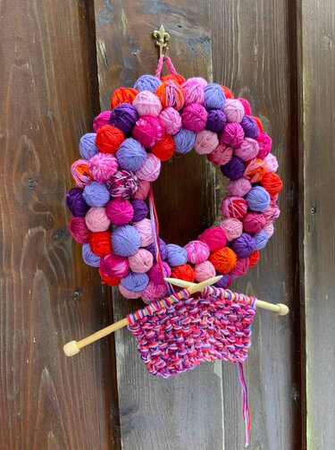 Strickkranz aus vielen bunten Garnknäueln mit Stricknadeln und kleinem Maschenstück an einer Holztür
