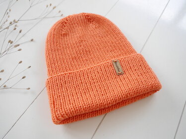 Orangene, gerippte Beanie-Mütze auf hellem Holz mit dezenten getrockneten Gräsern.