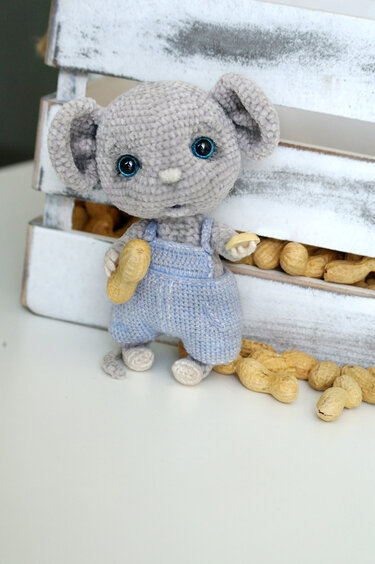 Gray crocheted amigurumi mouse wearing light-blue overalls and holding a peanut, posed by a whitewashed wooden crate of peanuts.