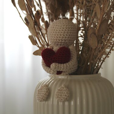 Beiges Amigurumi-Figürchen aus Häkelarbeit mit rotem Herz in einer weißen Vase mit Trockenblumen