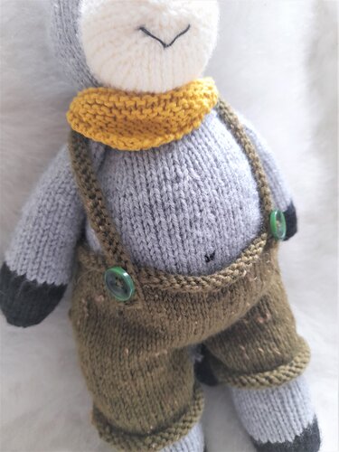 Gestricktes Amigurumi‑Schaf in brauner Latzhose mit grünen Knöpfen und gelbem Schal, Nahaufnahme.