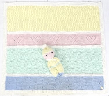 Draufsicht auf gestreifte Babydecke in Pastellfarben mit einer kleinen gestrickten Puppe; Details mit Herzmuster und Waffelstruktur sichtbar.