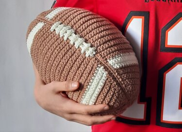 Hand hält ein gehäkeltes Football-Kissen in Braun und Weiß mit weißen Streifen