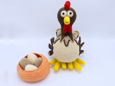 Gehäkeltes Amigurumi-Huhn in Braun-, Beige- und Gelbtönen neben einem orangefarbenen Korb mit gehäkelten Eiern auf hellem Hintergrund.