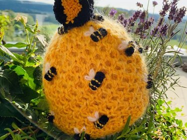 Gelb gestrickte Kindermütze mit kleinen Bienenapplikationen und schwarz-gelbem Pompon, zwischen Lavendel und grünen Blättern.