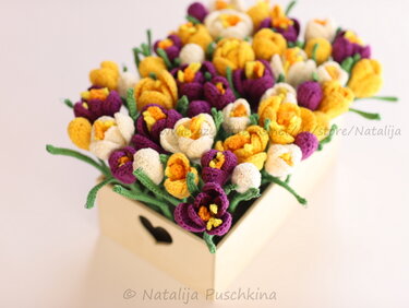 Top view of crocheted yellow, purple and white flowers with green stems in a light wooden box.