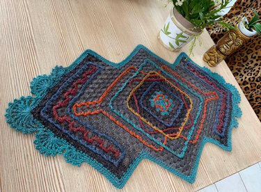 Gehäkelter Tischläufer in Anthrazit mit bunten Rauten- und Zickzackmustern und türkisfarbenem Muschelrand auf einem Holztisch.