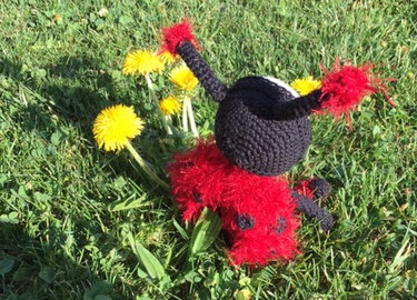Rotes und schwarzes gehäkeltes Amigurumi-Käferchen mit flauschigen Antennen auf Gras und Löwenzahn