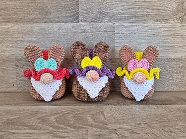 Drei gehäkelte Amigurumi-Osterwichtel mit braunen Hasenohren, weißem Bart, Rüschenkragen und bunten Schleifen vor Holz.