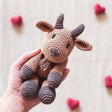 Gehäkeltes Amigurumi-Zicklein in einer Hand vor roten Herz-Dekoelementen