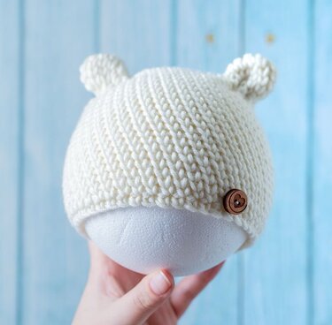 Nahaufnahme einer gehäkelten Babymütze in Creme mit Ohren und Holzknopf, gehalten von einer Hand.