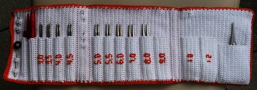 Aufgerolltes weißes Häkel-Etui mit orangefarbenem Rand, gefüllt mit Metall-Stricknadelspitzen und weiterem Strickzubehör.