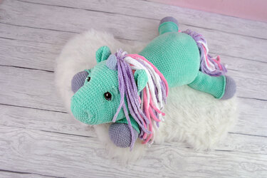 Teal crocheted amigurumi unicorn with purple, pink, and white mane lying on a white faux-fur rug.