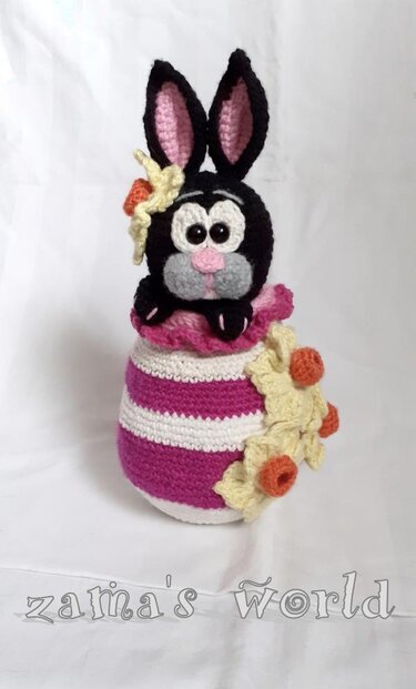 Gehäkelter schwarzer Hase mit rosa Innenohren, der aus einem weiß-rosa gestreiften Ei mit Blumen schaut.