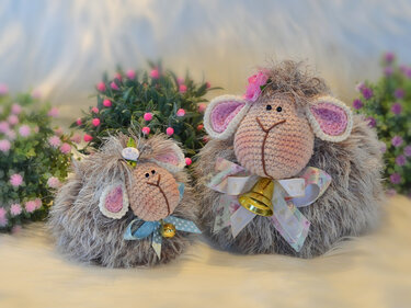 Zwei handgehäkelte Amigurumi-Schafe mit grauem flauschigem Körper, rosa Innenseiten an den Ohren, Schleifen und Glöckchen vor Blumen.
