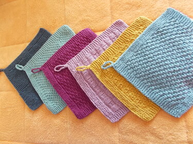 Reihe von sechs gestrickten Topflappen in verschiedenen Farben mit Aufhängeschlaufen auf orangefarbener Unterlage