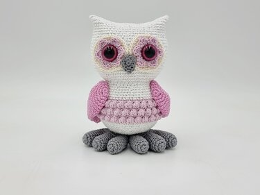 Gehäkelte Amigurumi-Eule mit rosa Flügeln, grauen Füßen und weißem Körper vor neutralem Hintergrund
