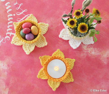Three yellow and white crocheted flower bowls on a pink surface; one holds pastel eggs, one a small bouquet, and one a lit tea light.