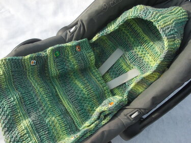 Grün melierter gestrickter Baby-Fußsack mit Kapuze und Knöpfen in einer Babyschale