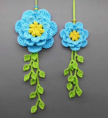 Two crocheted hanging decorations, large and small, each with a blue layered flower, yellow center, and trailing green leaf vines.