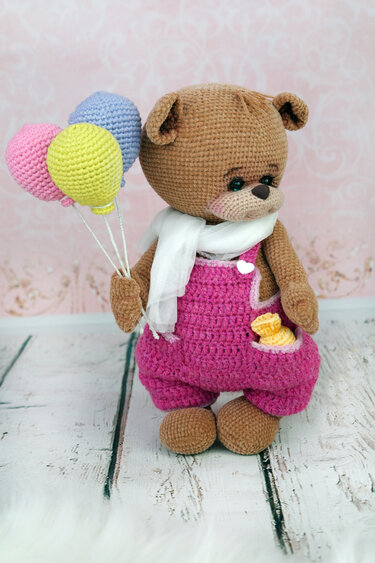 Brown crocheted teddy bear wearing pink overalls and a white scarf, holding three crocheted balloons.