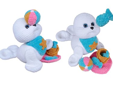 Two white crocheted amigurumi seals with a colorful beach ball, sand bucket, and fish accessory.