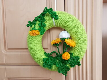 Hellgrüner gehäkelter Türkranz mit grünen Blättern, gelben Löwenzahnblüten und weißer Pusteblume an einer Holztür