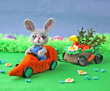 Gray crocheted amigurumi rabbit in blue overalls driving an orange carrot car with button wheels, pulling a small trailer filled with crochet carrots and flowers on a green backdrop.