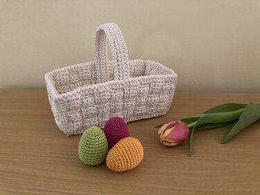 Beiges gehäkeltes rechteckiges Körbchen mit Henkel, davor drei gehäkelte Eier (grün, violett, orange) und eine Tulpe auf einem Holztisch