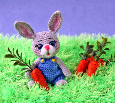 Gehäkelter grauer Amigurumi-Hase in blauer Latzhose mit Karotte im grünen Gras