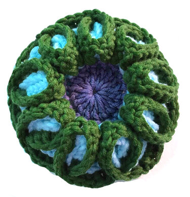 Rundes gehäkeltes Blumenmotiv mit grünen Schlaufen, hellblauen Einsätzen und violettem Zentrum auf hellem Hintergrund.