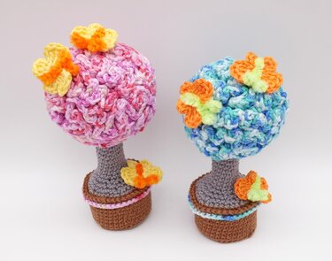Zwei gehäkelte Blumenbäume in Töpfen, links rosa/pastell mit gelb-orange Blüten, rechts blau/grün mit orange-gelben Blüten
