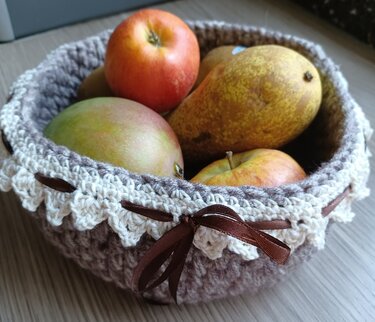 Gehäkelter Korb in Braun und Beige mit cremefarbener Spitze und braunem Satinband, gefüllt mit Äpfeln, Mango und Birne.