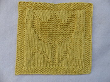 Quadratischer, gelber gestrickter Untersetzer mit Tulpenmotiv in der Mitte.