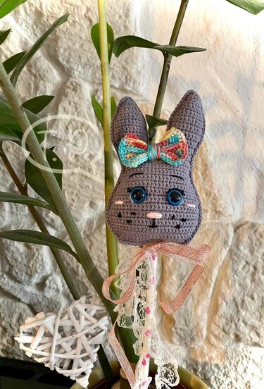 Gehäkelter Hasenkopf mit bunter Schleife und Spitzenband als Pflanzenstecker im Blumentopf