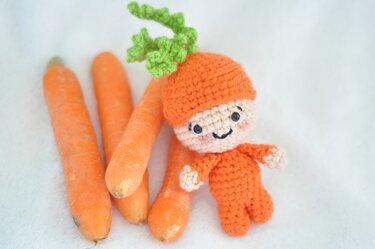Kleines orange‑grünes Amigurumi in Karottenform liegt neben drei Möhren auf weißem Tuch